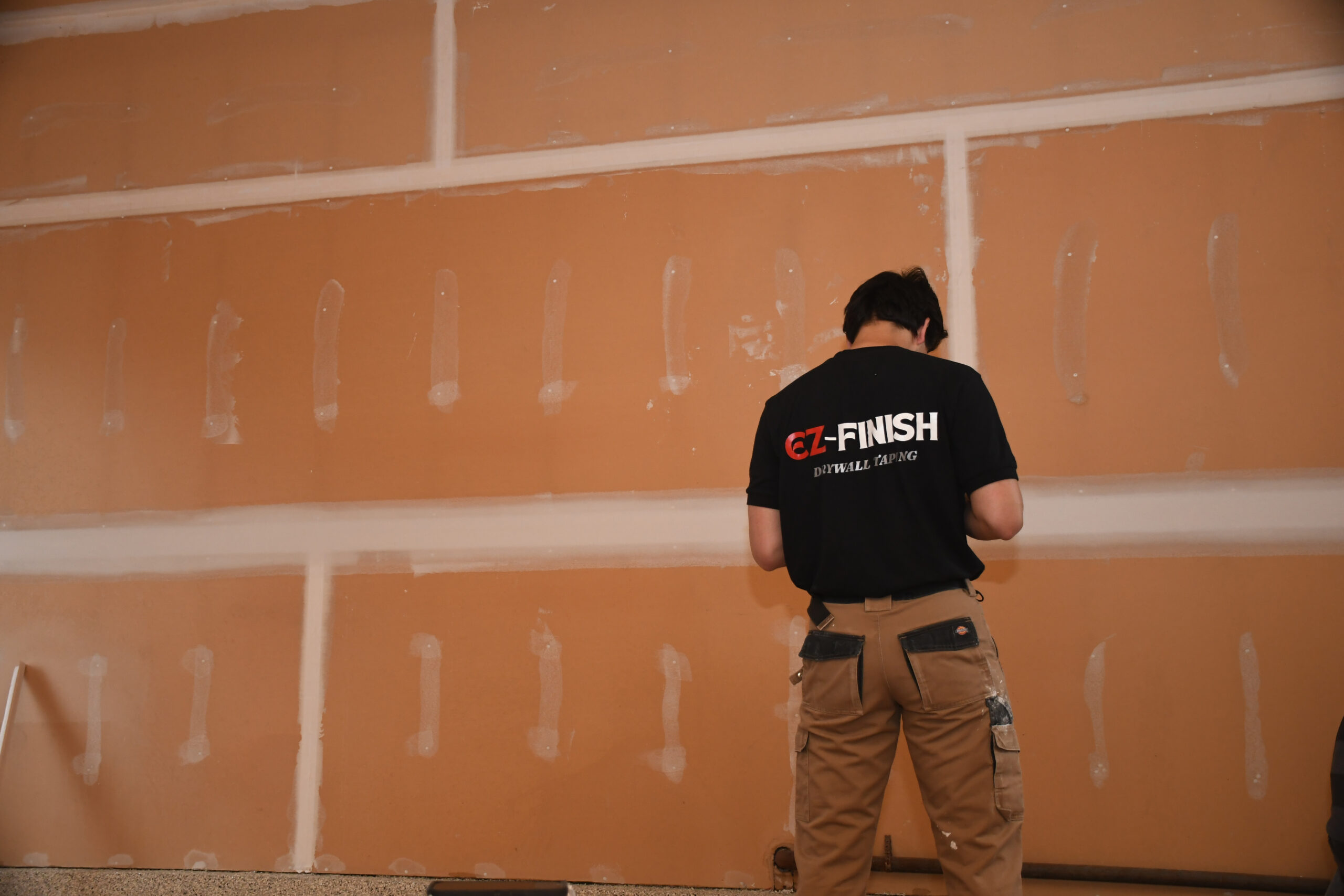 Not for the graphics, but for the storytelling. Worker with back to camera installing drywall in an unfinished orange wall, wearing a black CZ-FINISH shirt and cargo pants.