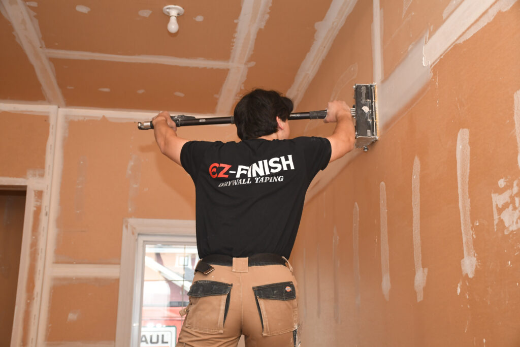 Worker in a CZ-Finish Drywall Taping shirt uses a long pole tool to install drywall on an unfinished orange wall.