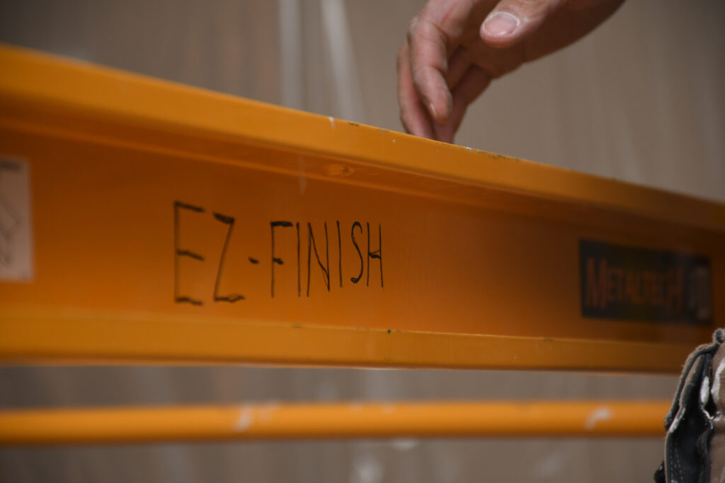 Close-up of a hand reaching toward the edge of an orange metal shelf in a workshop, with black handwritten text on the side.
