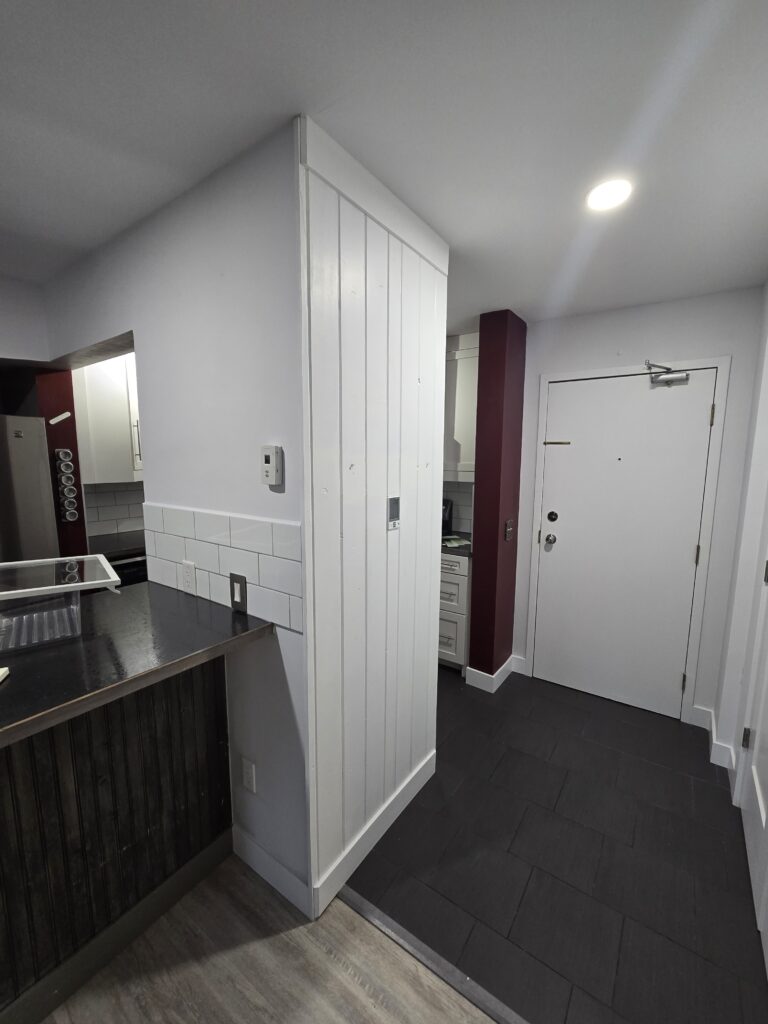 Modern kitchen area featuring white paneled wall, dark countertop, and a white door beside a burgundy pillar/support column.