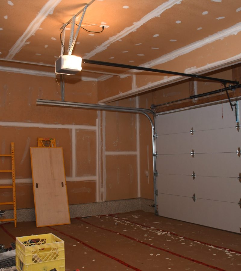 Garage under renovation with taped drywall seams, exposed wiring, and a hanging work light; ladder and tools on the floor.